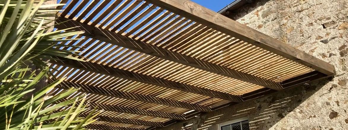 Pergola en bois à lames ajourées posée contre une façade en pierre, sous un ciel bleu